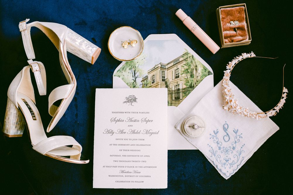 Wedding accessories including white heels, invitation, pearl necklace, and embroidered handkerchief arranged on dark surface.
