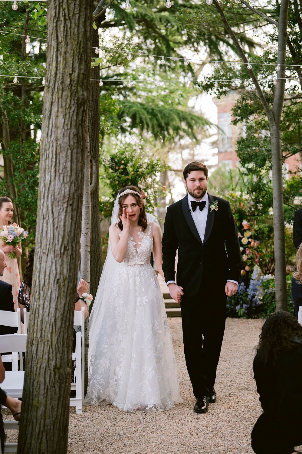 Wedding party walking through a grove of tall cedar trees on a gravel path during an outdoor ceremony.