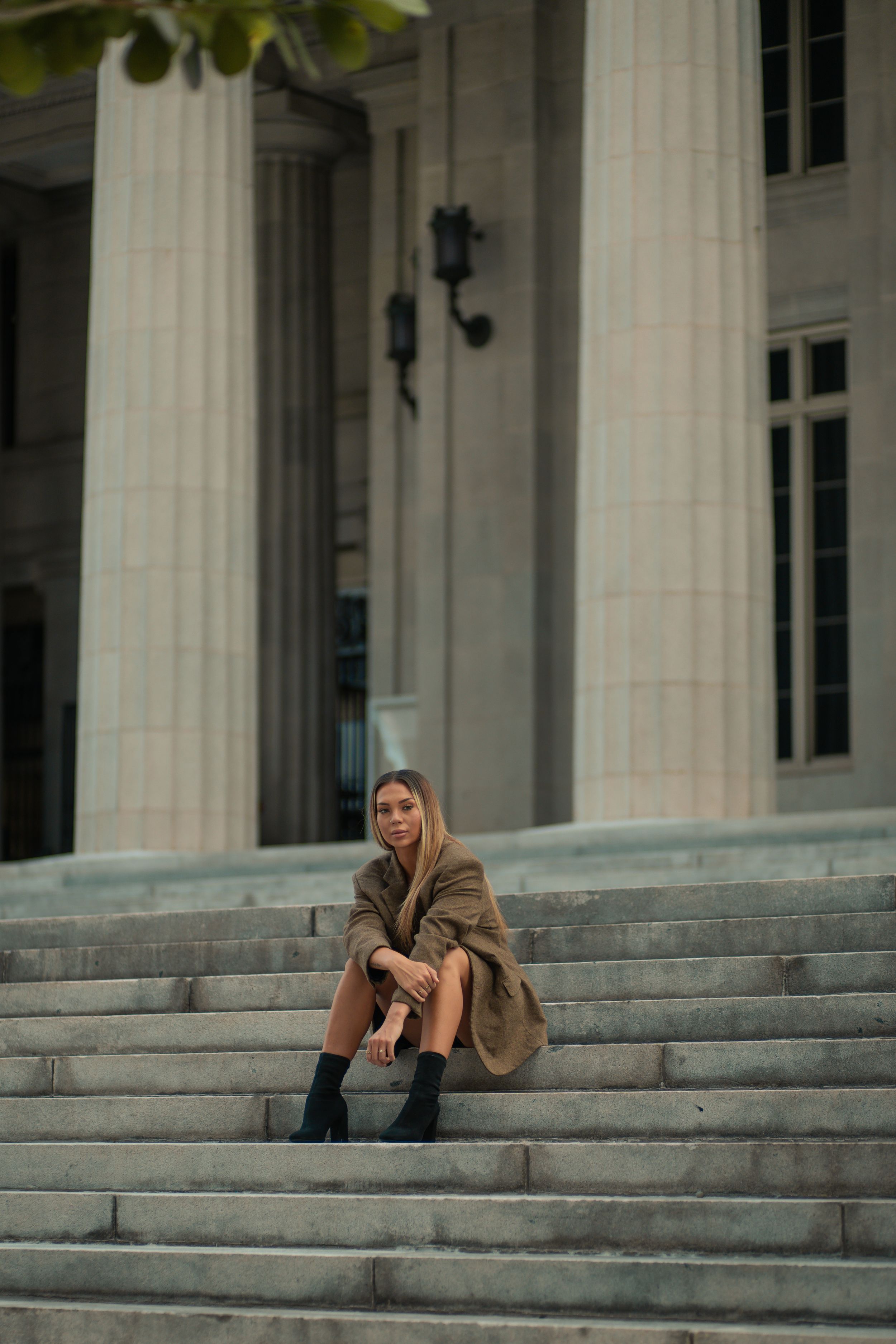 Close-up of woman in brown jacket leaning forward on courthouse stairs, Miami fashion editorial portrait, cinematic photography style.