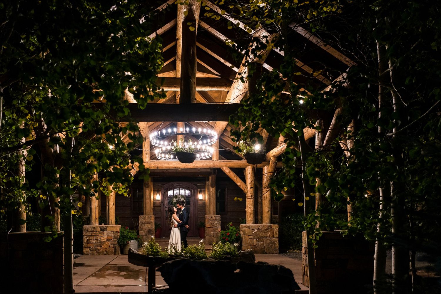 Rustic wooden pavilion surrounded by trees and illuminated with warm lighting at night.