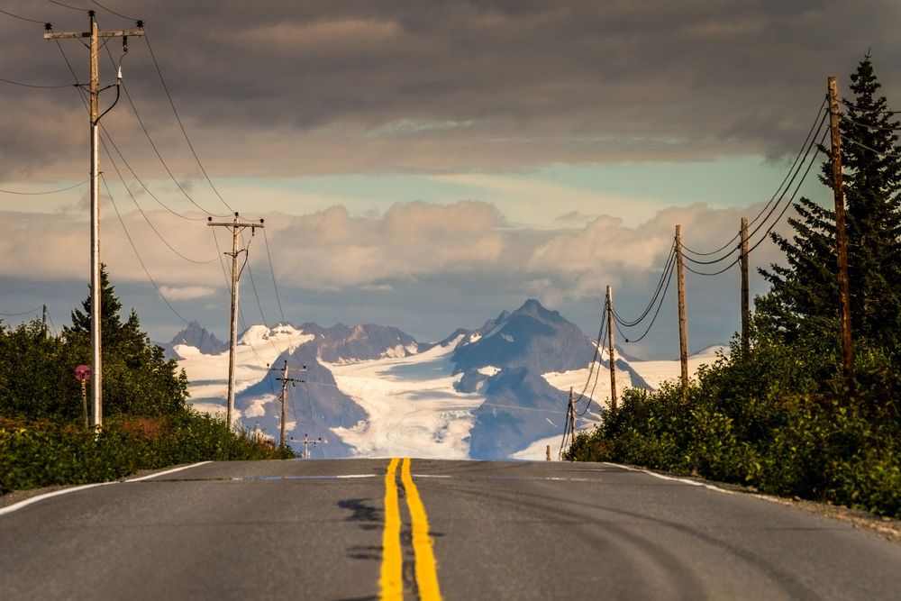 A dramatic mountain highway leads into snow-capped peaks with power lines and evergreen trees along the roadside.