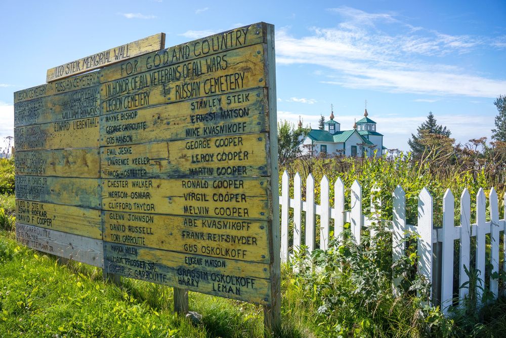 Old-style directional signage mounted on wooden post indicates distances to destinations against a bright blue sky and foliage.