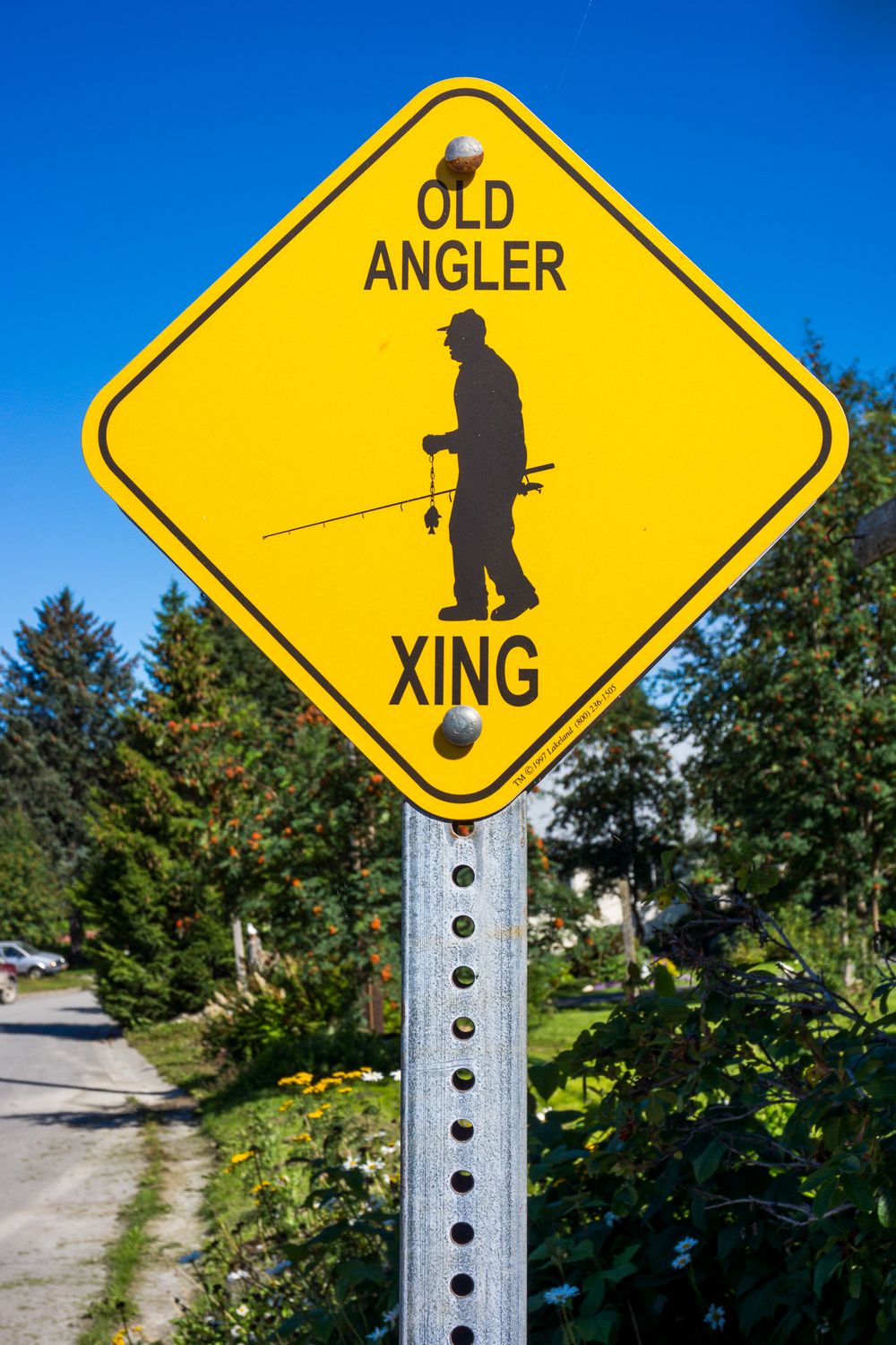 Yellow diamond-shaped road sign showing a silhouette of an angler indicates a fishing area crossing ahead on a sunny day.