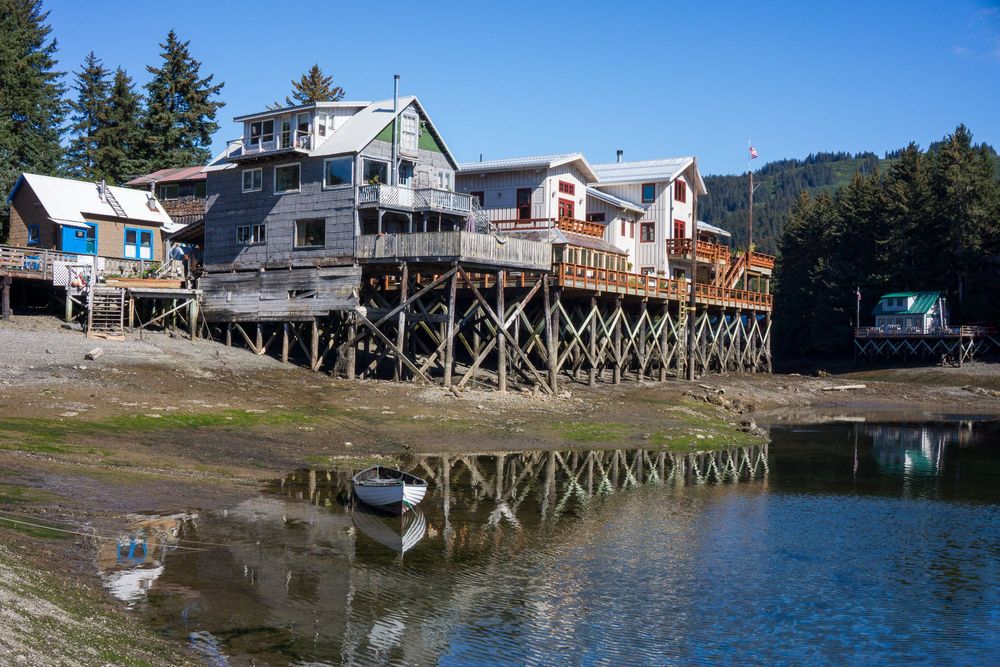 Coastal homes on wooden stilts reflect in calm waters at low tide with evergreen forest in the background.