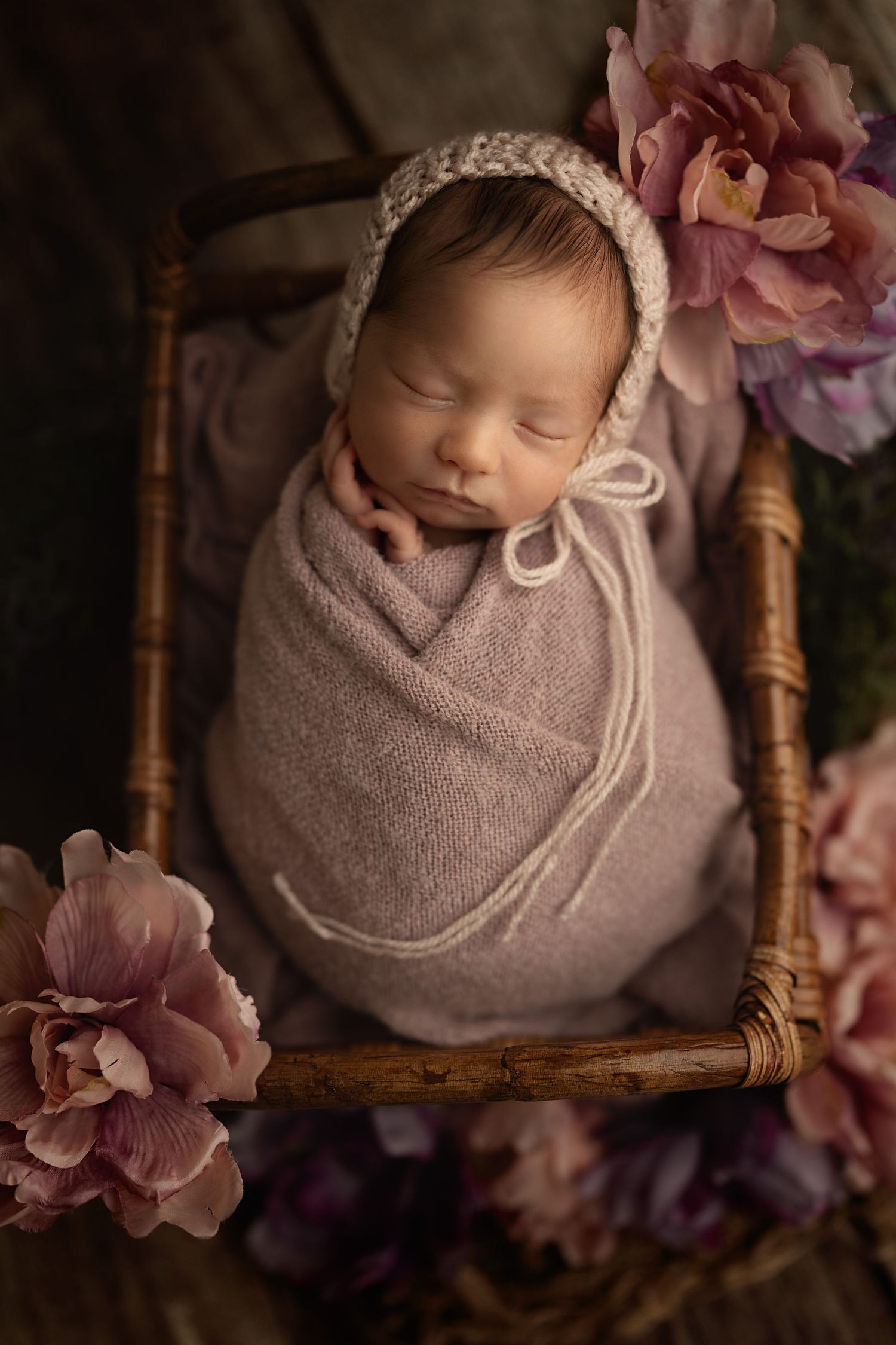 Artistic portrait of sleeping newborn in pink wrap with floral accents in vintage wooden basket.