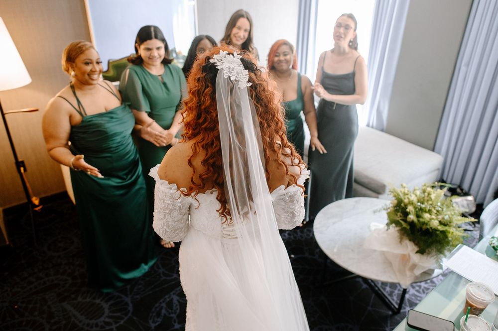 Bride getting dressed in a white bridal suite with bridesmaids in emerald green dresses assisting with final touches.