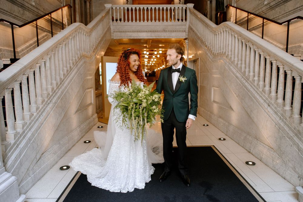 A couple in formal wedding attire standing on an elegant marble staircase with white railings during their celebration.