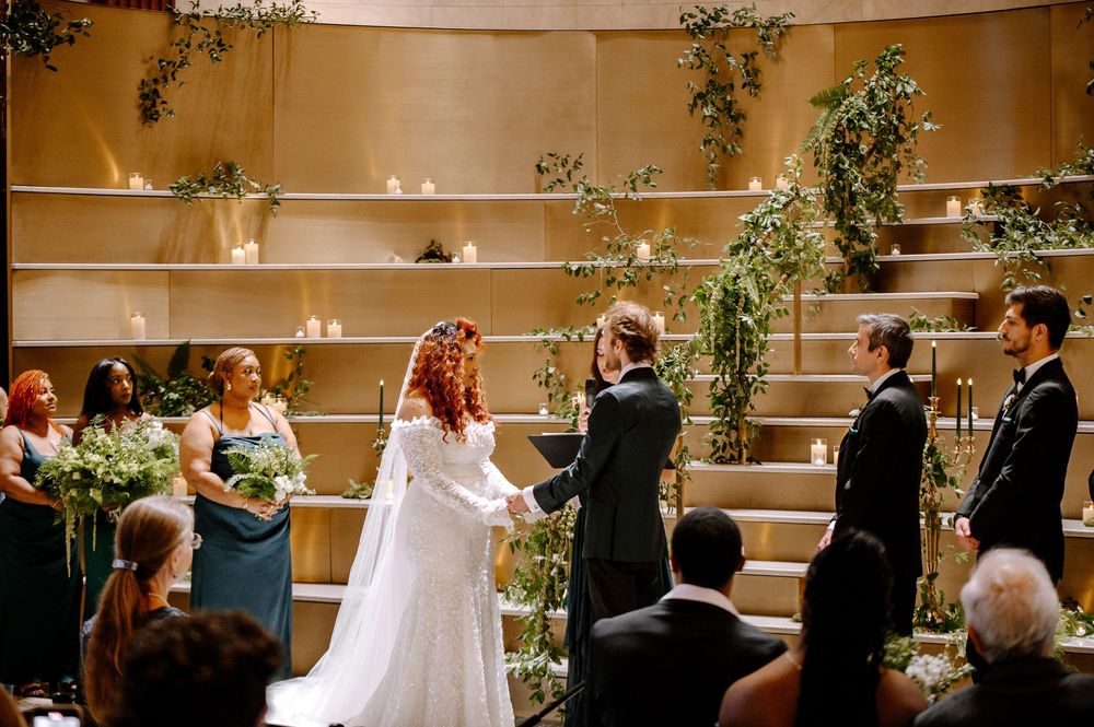 Indoor wedding ceremony scene with guests seated in curved rows watching a couple exchange vows at the altar.