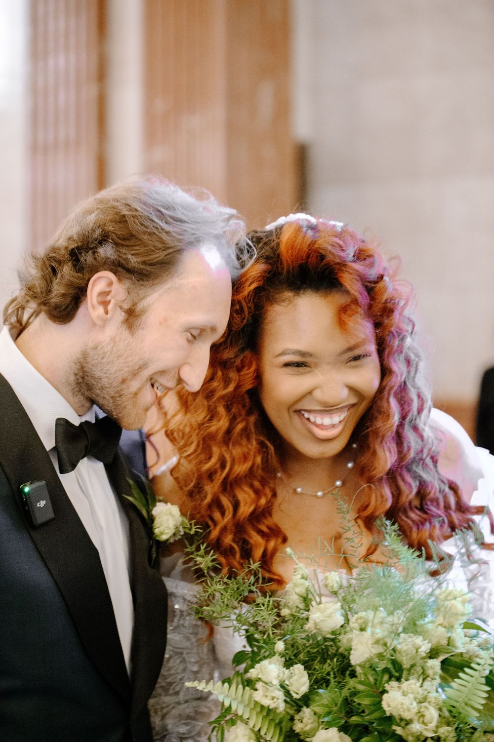 Joyful wedding moment showing a bride with vibrant curly orange hair holding white bouquet next to groom in tuxedo.