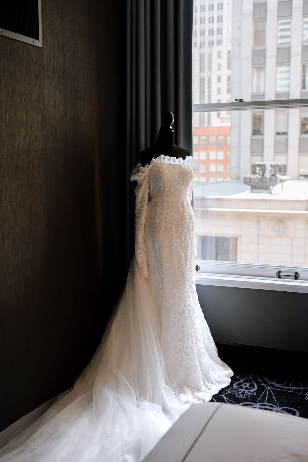 A white lace wedding dress hangs elegantly by a window in a hotel room with city buildings visible outside.