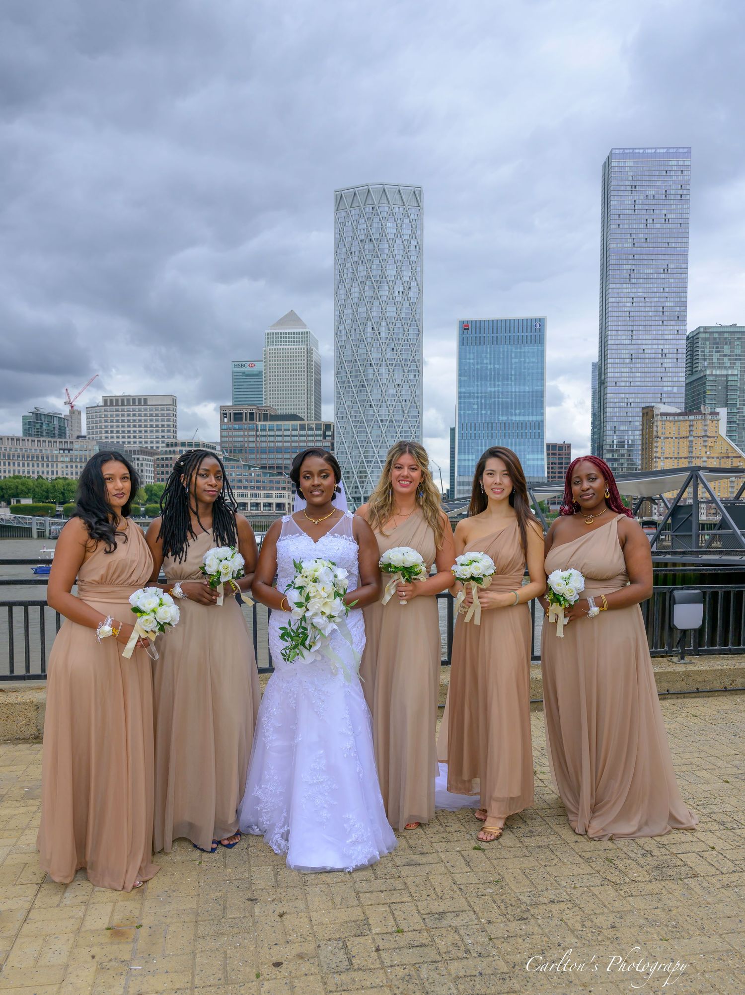 A bridal party in tan bridesmaid dresses and a white wedding gown pose against a backdrop of modern city buildings.