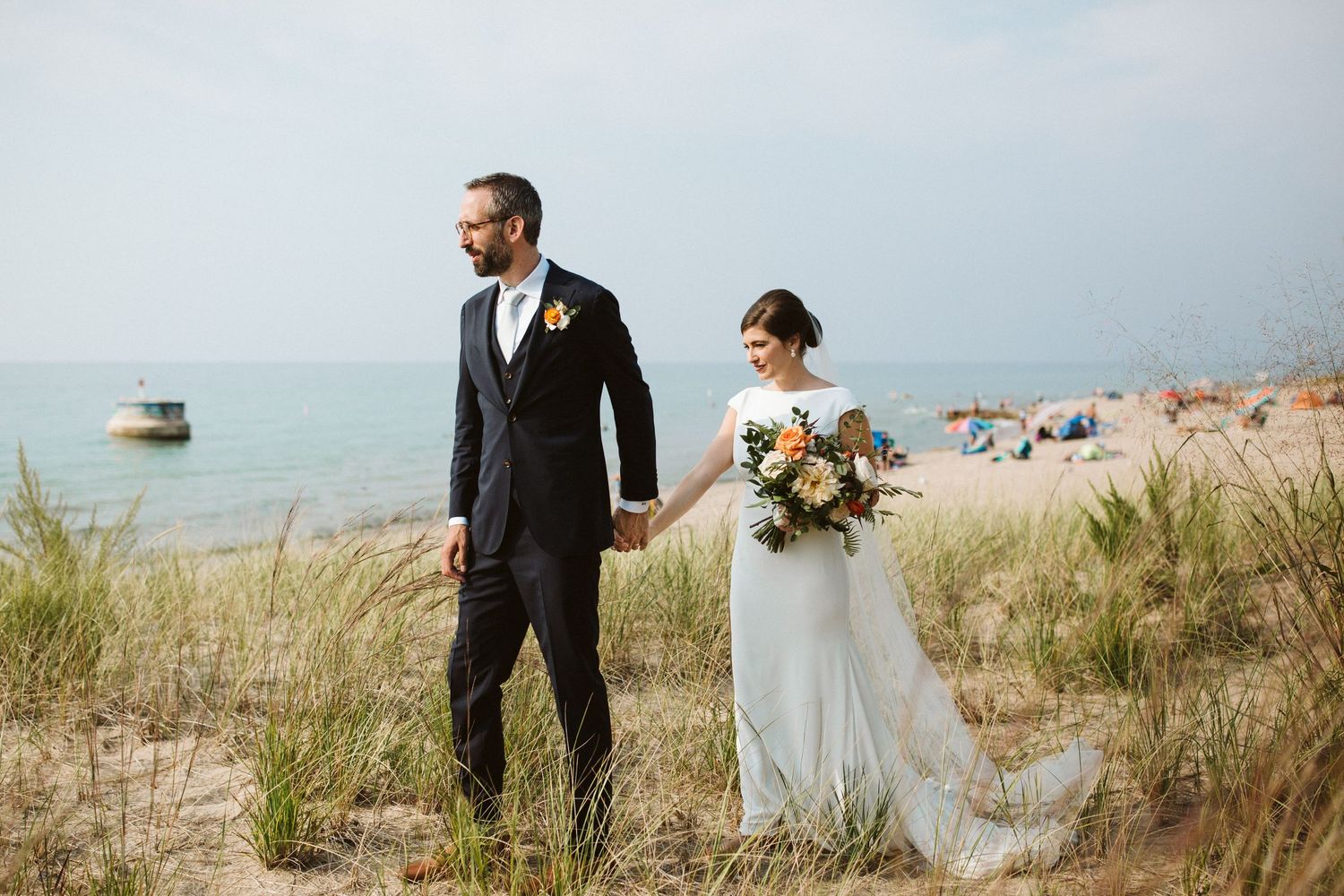 Wedding couple poses together on sandy beach dunes with lake in background.