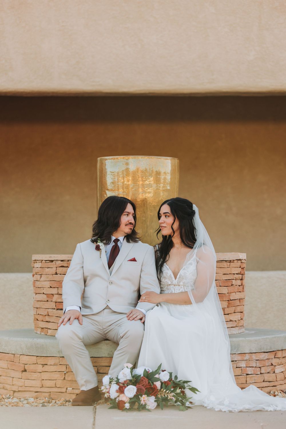 Happy wedding couple poses lovingly against stone wall in creamy beige outfits during intimate outdoor ceremony.