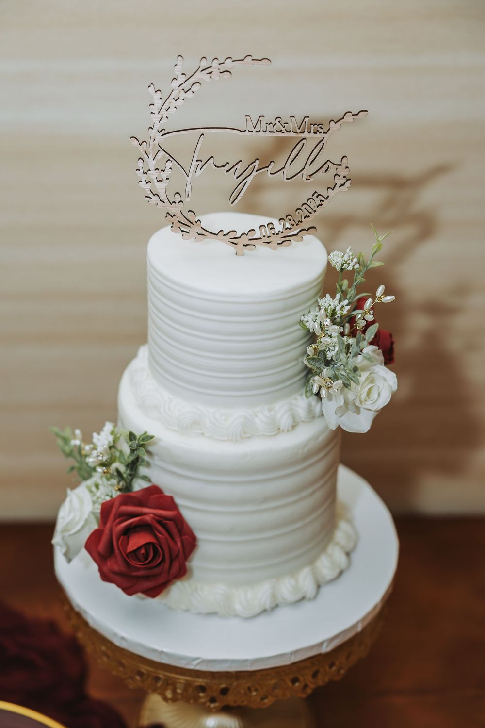 White tiered wedding cake decorated with red roses and delicate branch topper displayed on wooden surface.