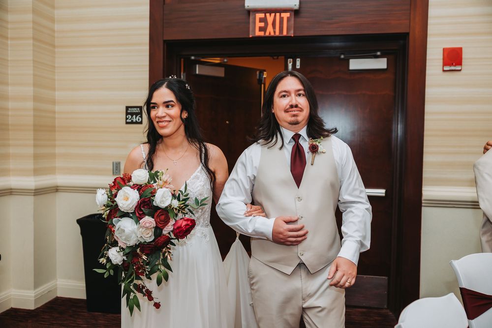 Two people in wedding attire share a laugh as they stand by a wooden exit door in a light-colored hallway.