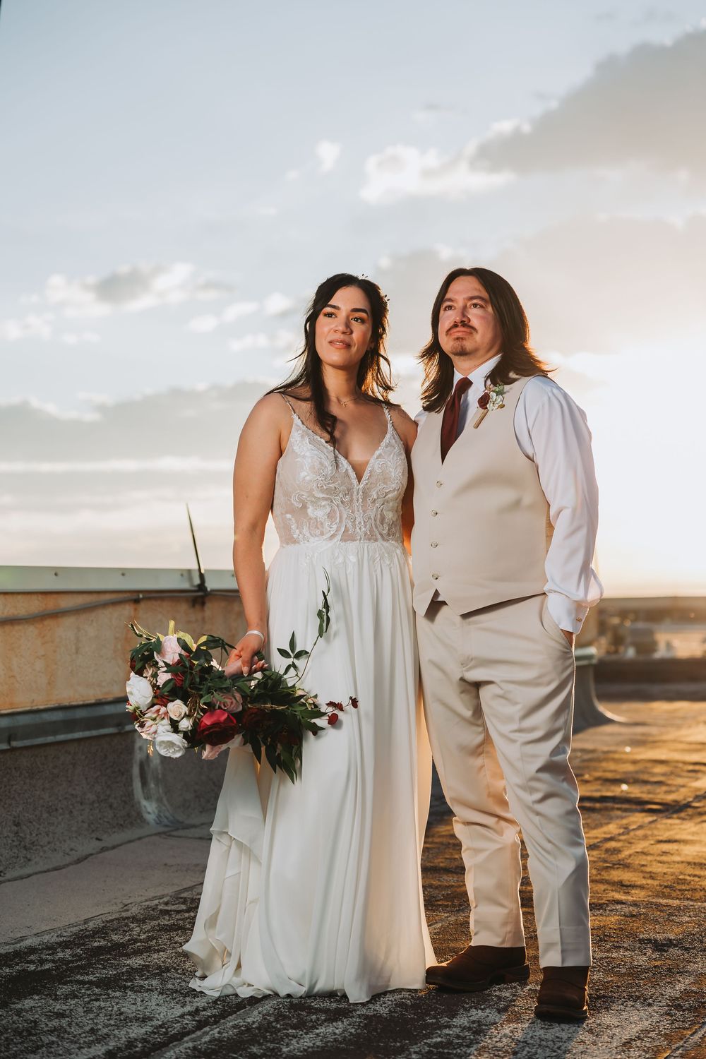 A wedding couple poses together at sunset on a rooftop with a dramatic sky backdrop and bouquet of flowers.