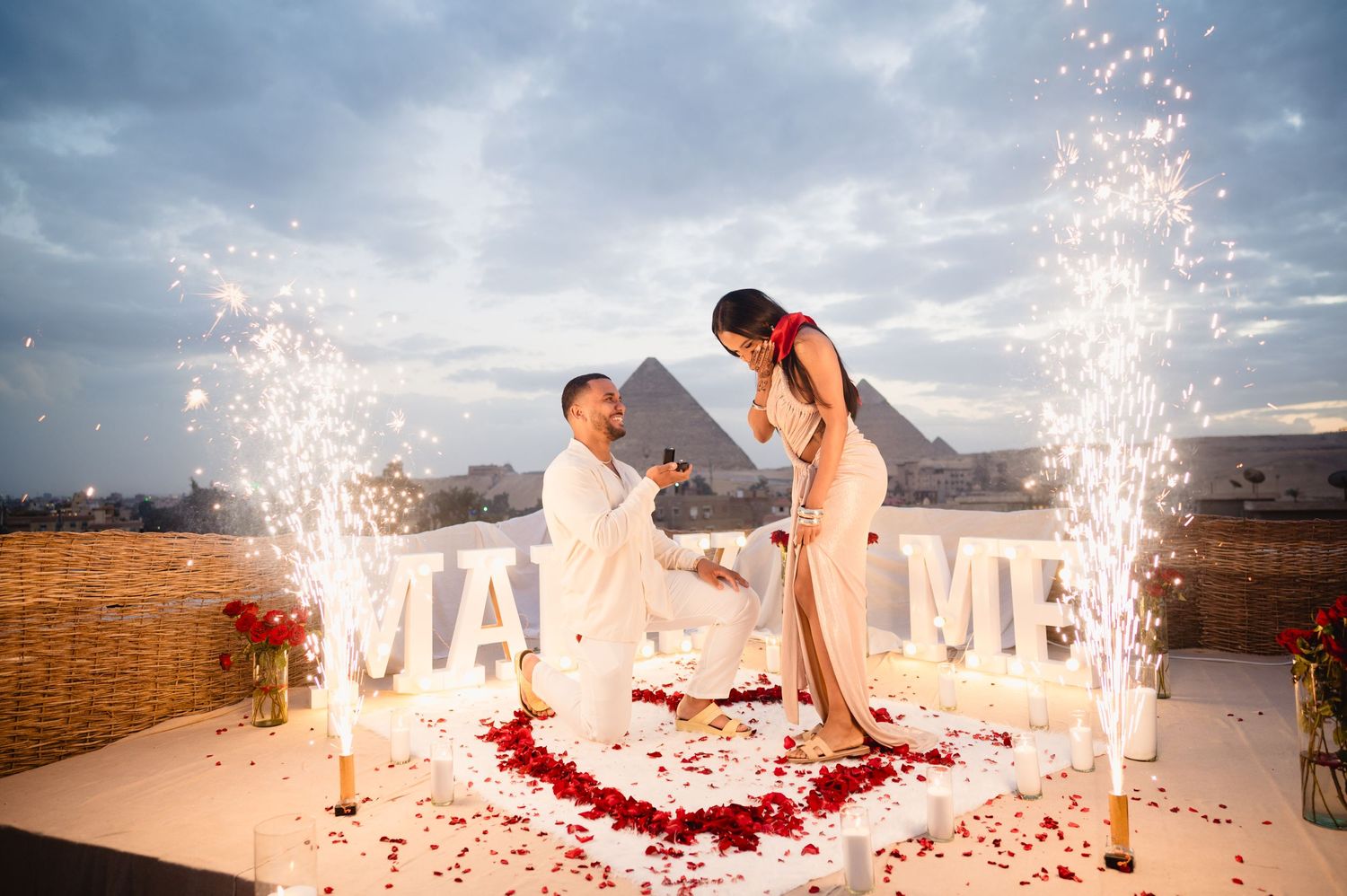 A romantic proposal scene with sparklers and rose petals against a dramatic sunset sky.