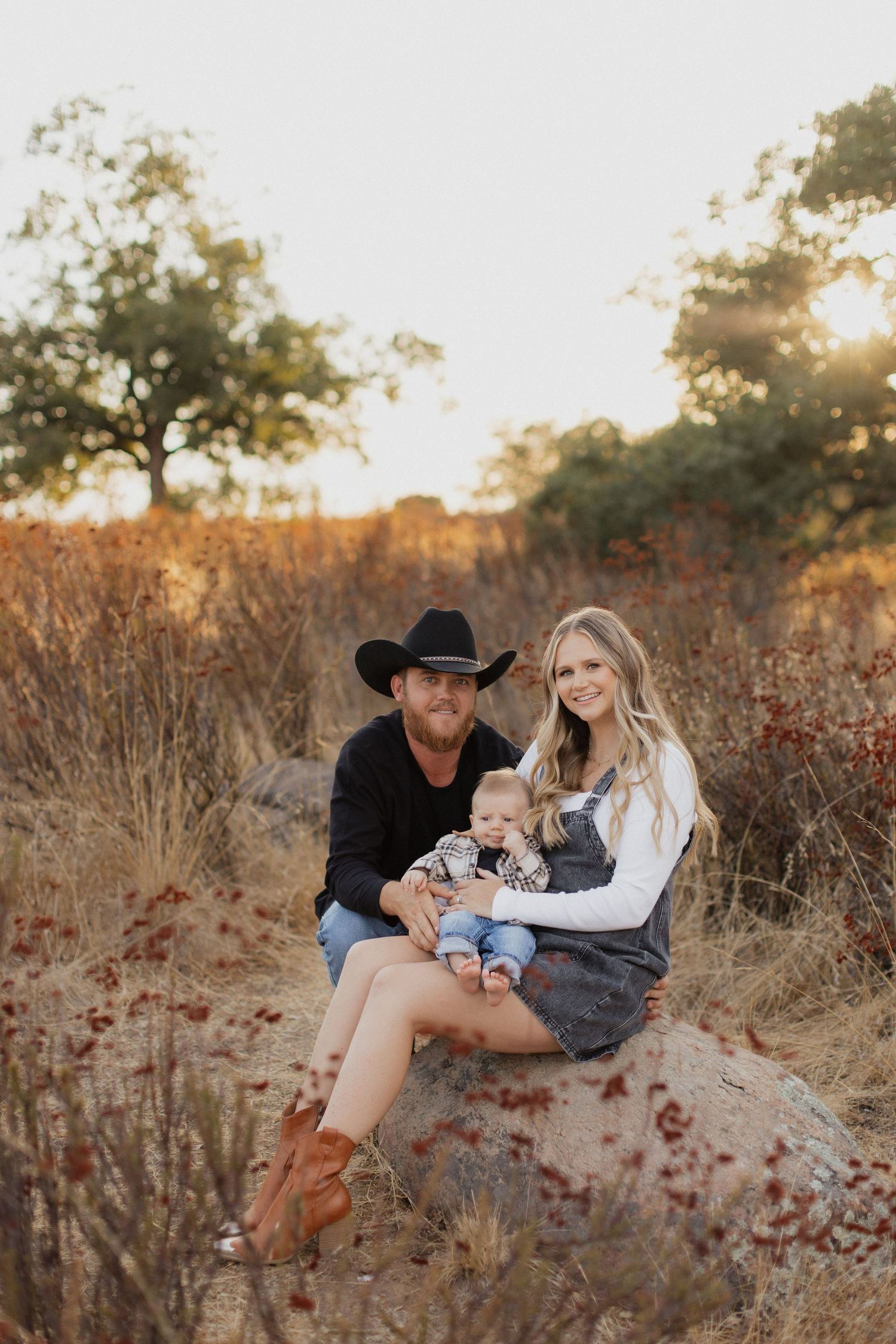 A couple sits together in a golden field at sunset amidst rustic fall foliage.