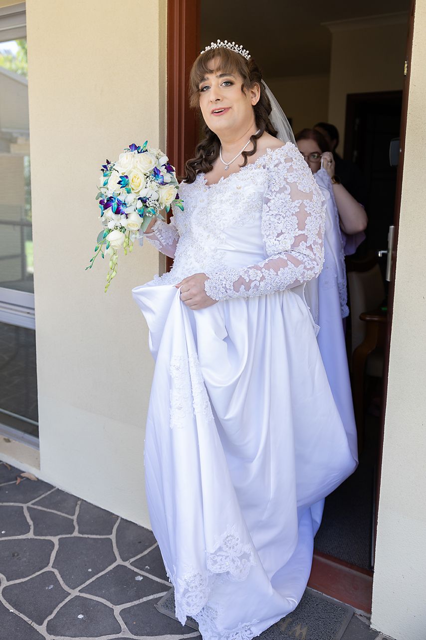 A bride in an elegant white lace wedding gown carries a blue and white bouquet while entering a doorway.