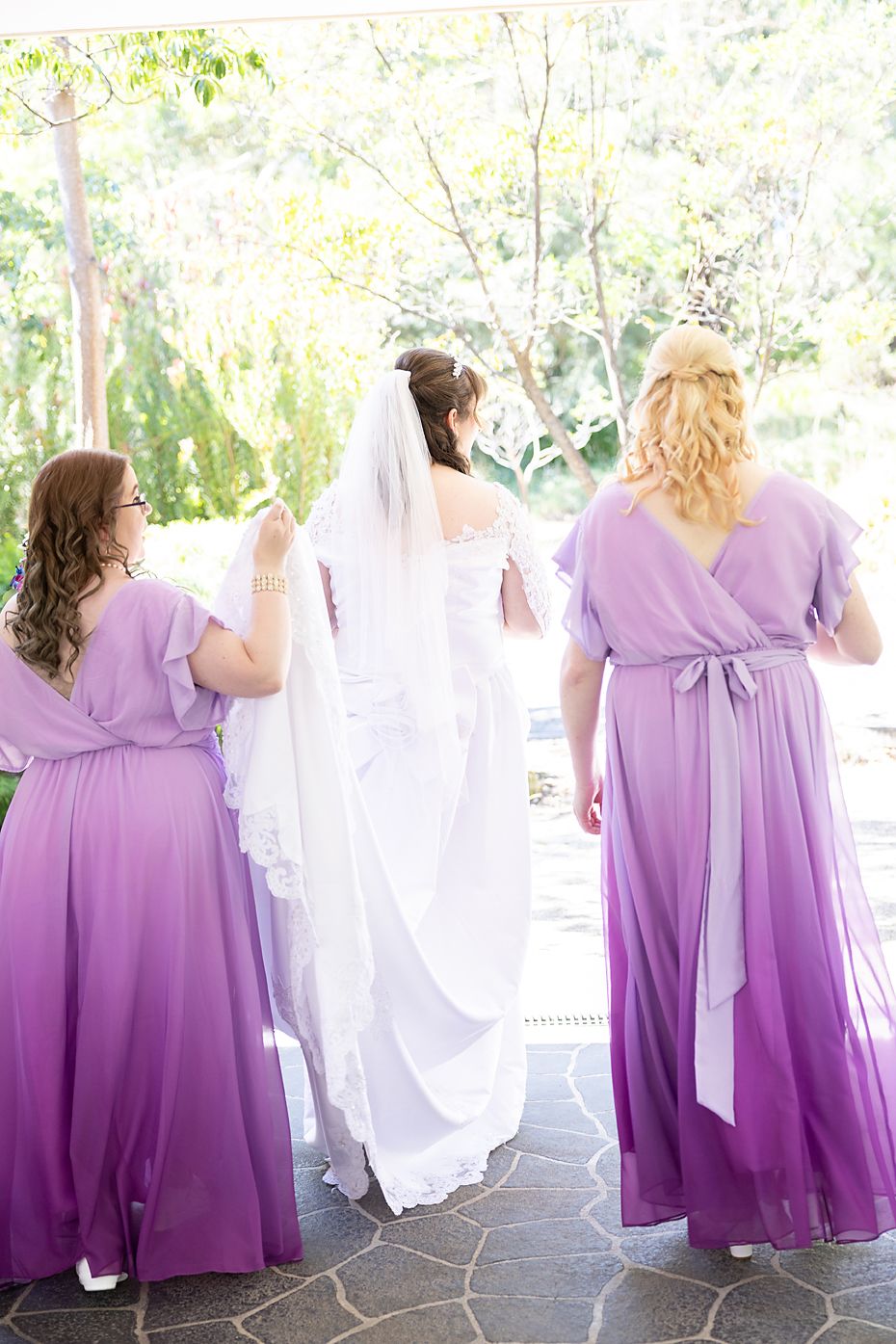 A line of bridesmaids wearing flowing purple ombre dresses viewed from behind at an outdoor wedding venue.