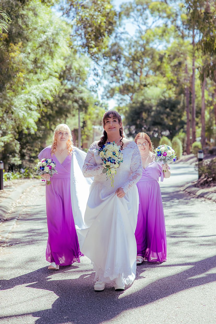 Bridesmaids in flowing purple dresses pose playfully on a tree-lined path with sunlight filtering through leaves.