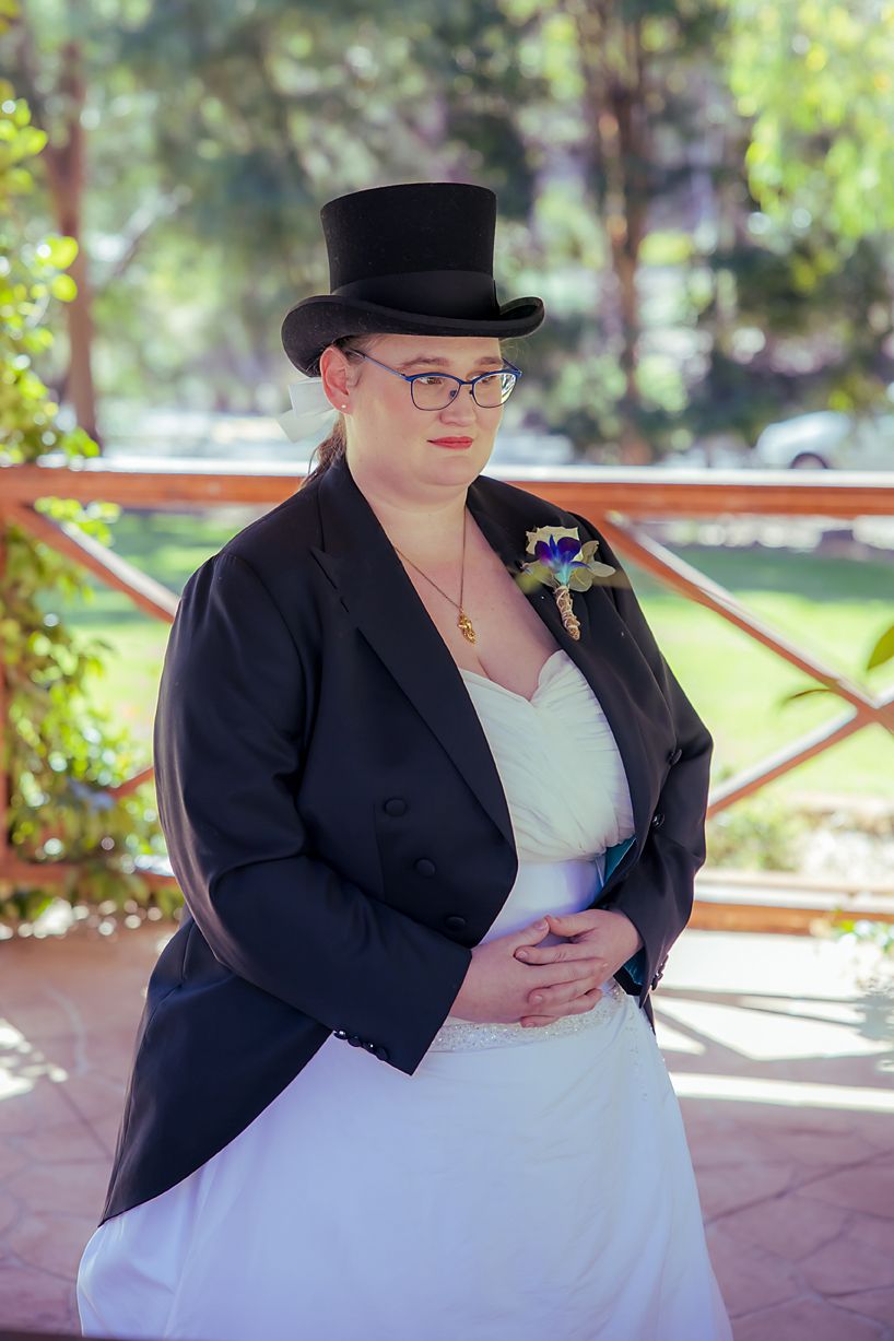 Person in black blazer and white dress sits outdoors checking phone while wearing glasses and bowler hat.