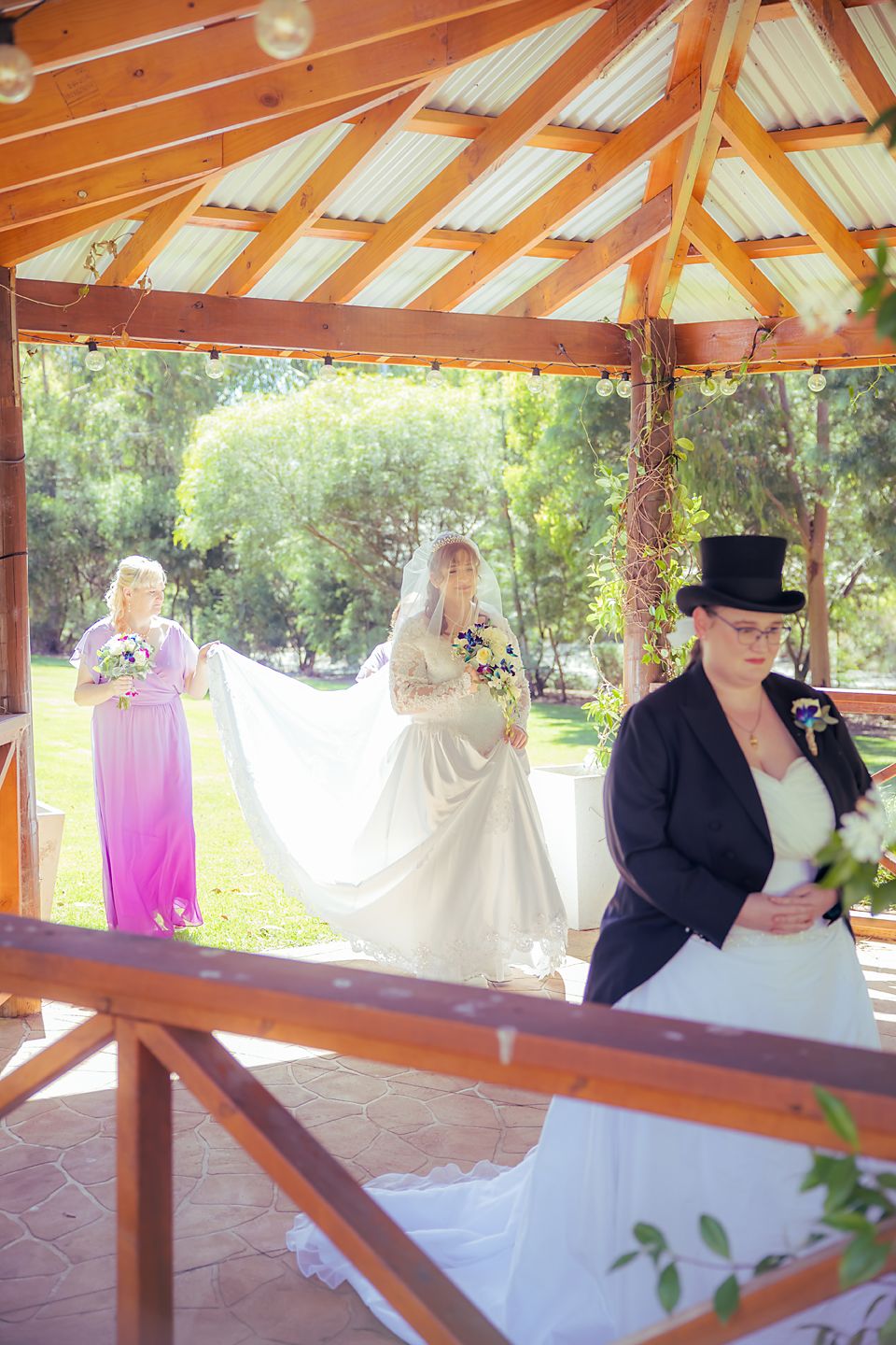 A couple embraces under a wooden gazebo structure with white and purple decorative elements in a garden setting.