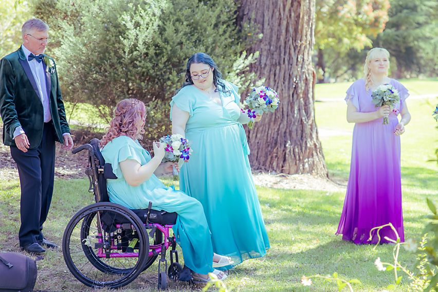 Wedding party gathered outdoors in teal and purple dresses poses under tall trees during summer celebration.