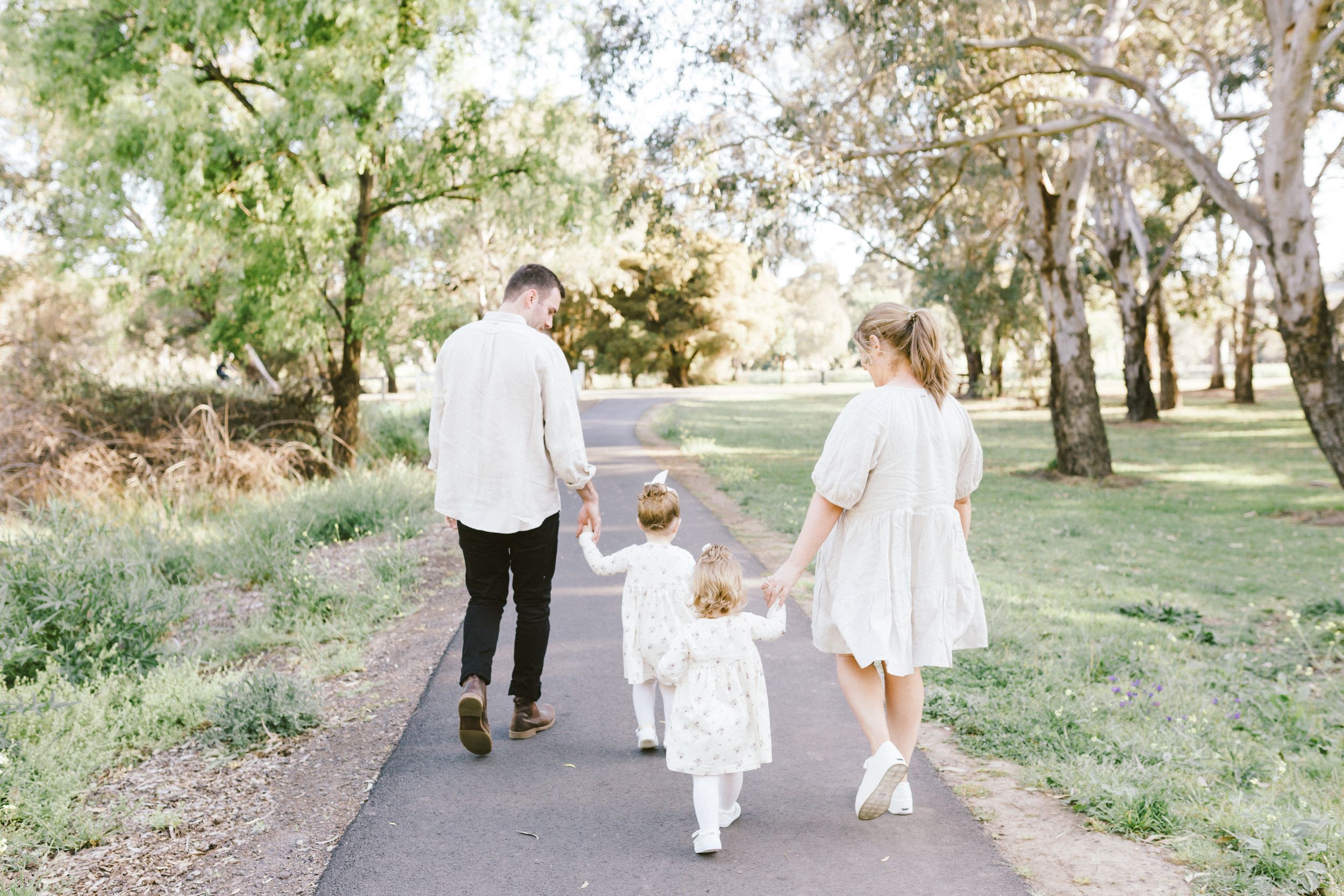 Family wearing white clothing walks together through a lush garden path during a sunny outdoor photo session.