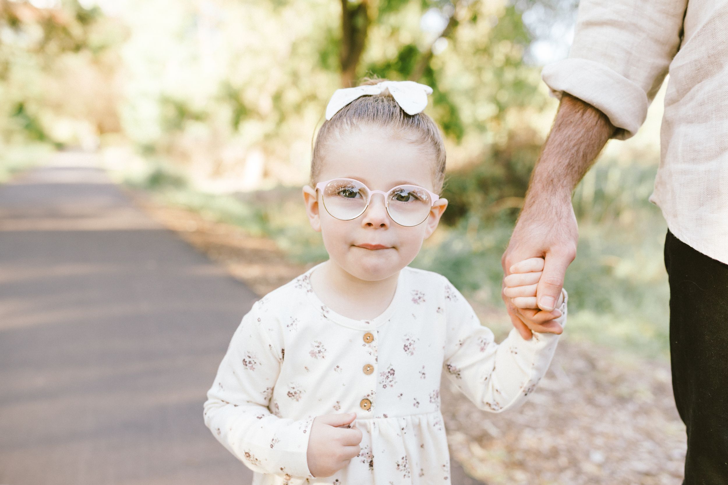 Small child wearing glasses and white sweater holds hand with adult while standing on dirt path outdoors.