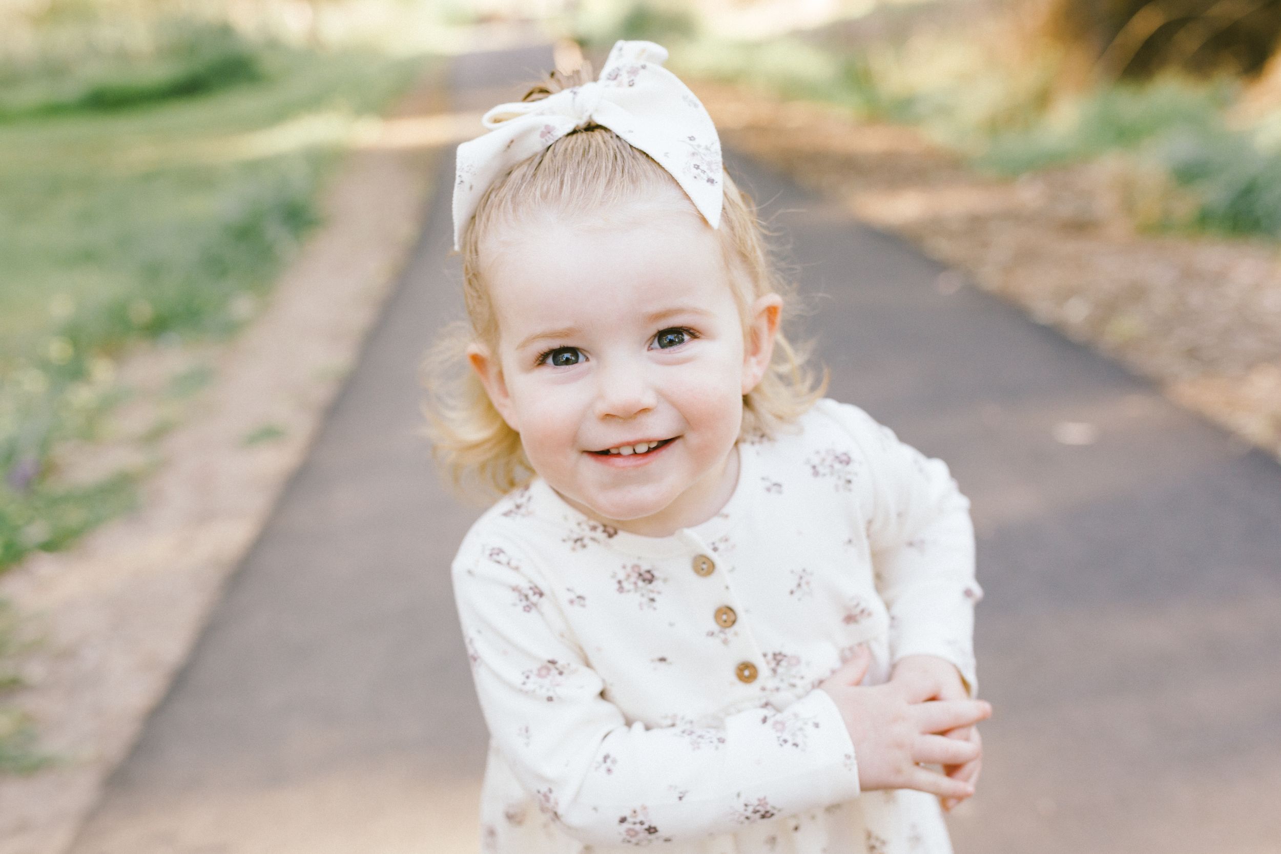 Little one wearing white lace dress with bow stands playfully on outdoor path in warm sunlight.