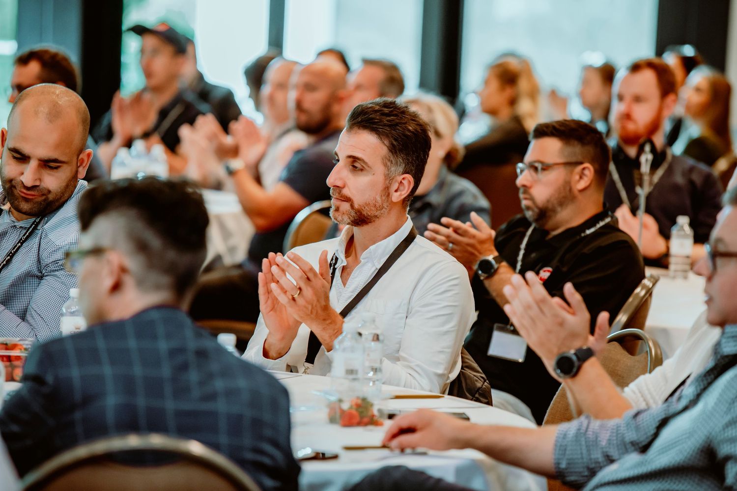 A diverse crowd of event attendees sitting at round tables and applauding during a business conference or presentation.