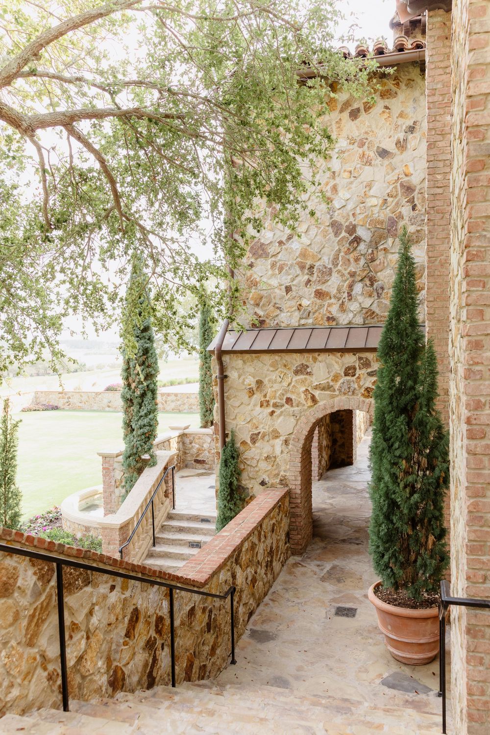 Rustic stone patio with tall potted cypress trees and wrought iron railings along a Mediterranean style veranda.