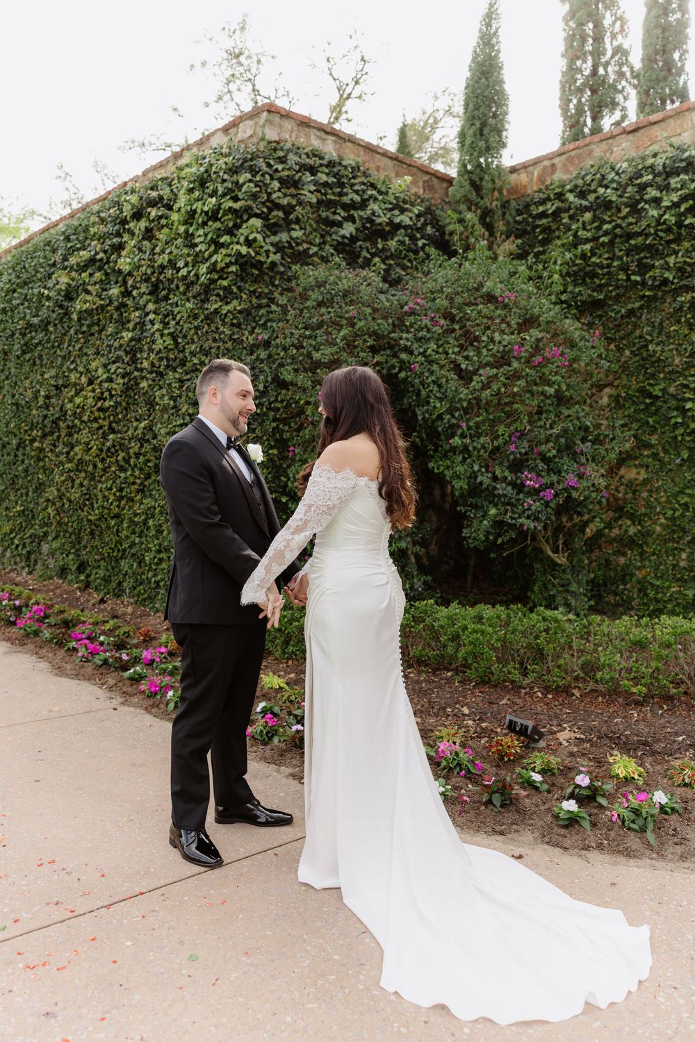 A couple share an intimate moment in formal wedding attire against a flowering garden backdrop with lush hedges.