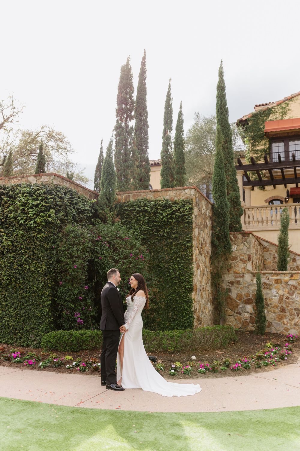 A couple shares an intimate moment in front of a stone wall and cypress trees in an elegant garden setting.