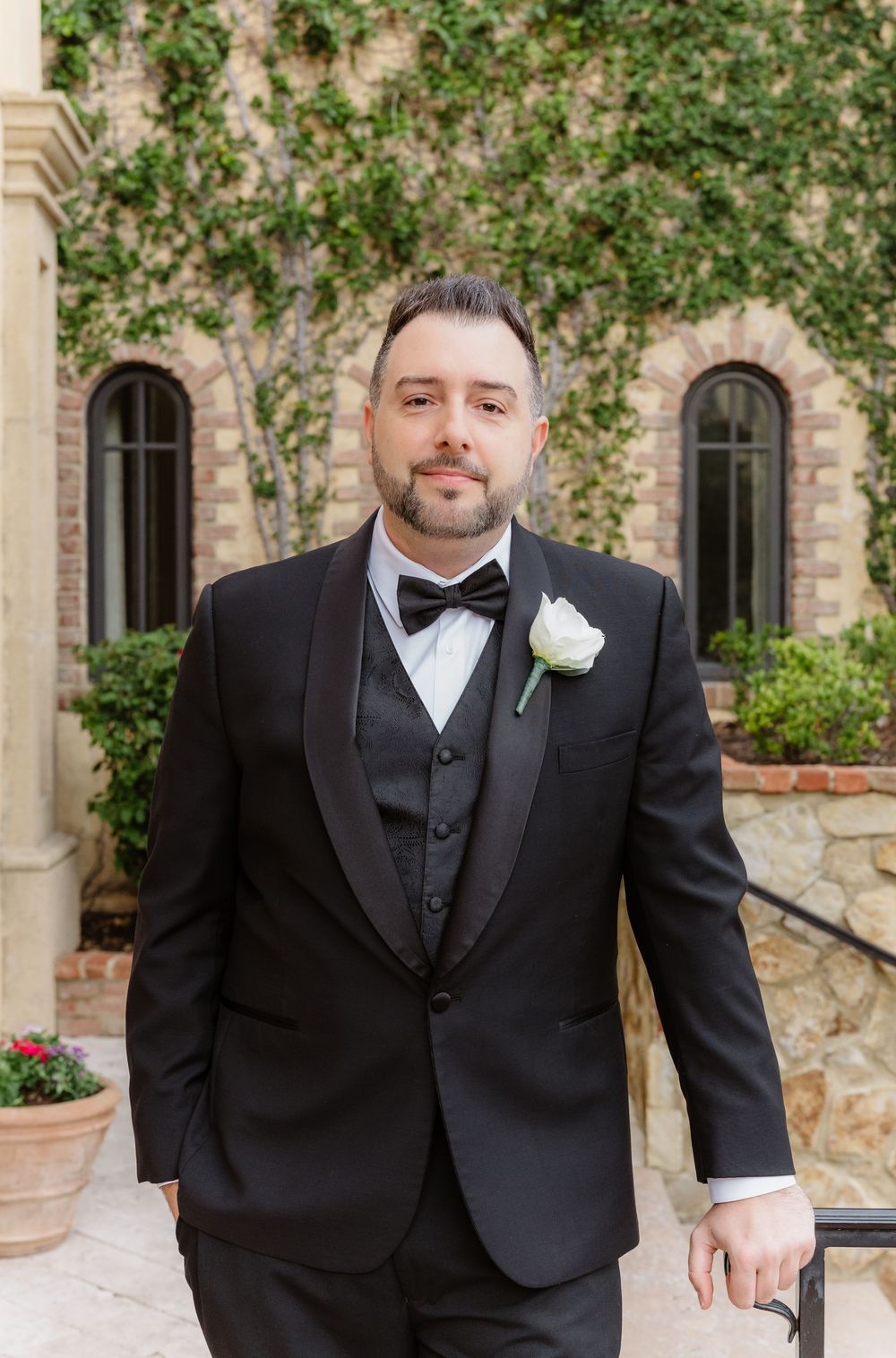 Three men in black tuxedos and bow ties pose outdoors in front of a stone building with climbing greenery.