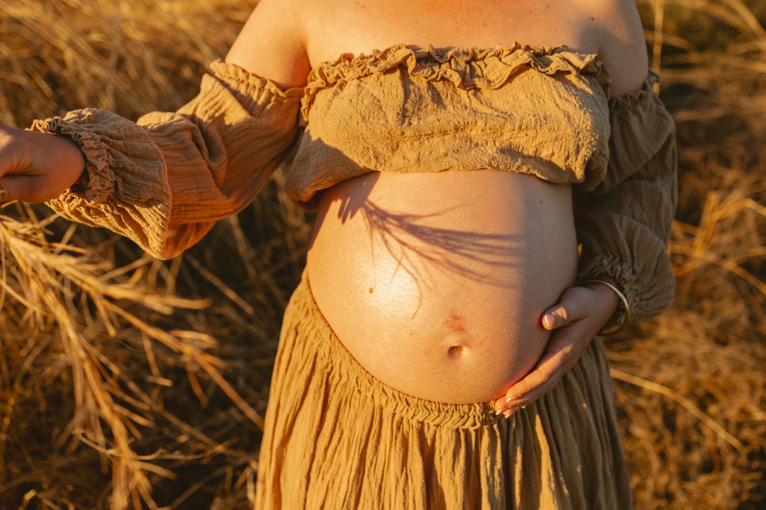 Silhouette of an expectant mother in a flowing dress against a golden field at sunset.