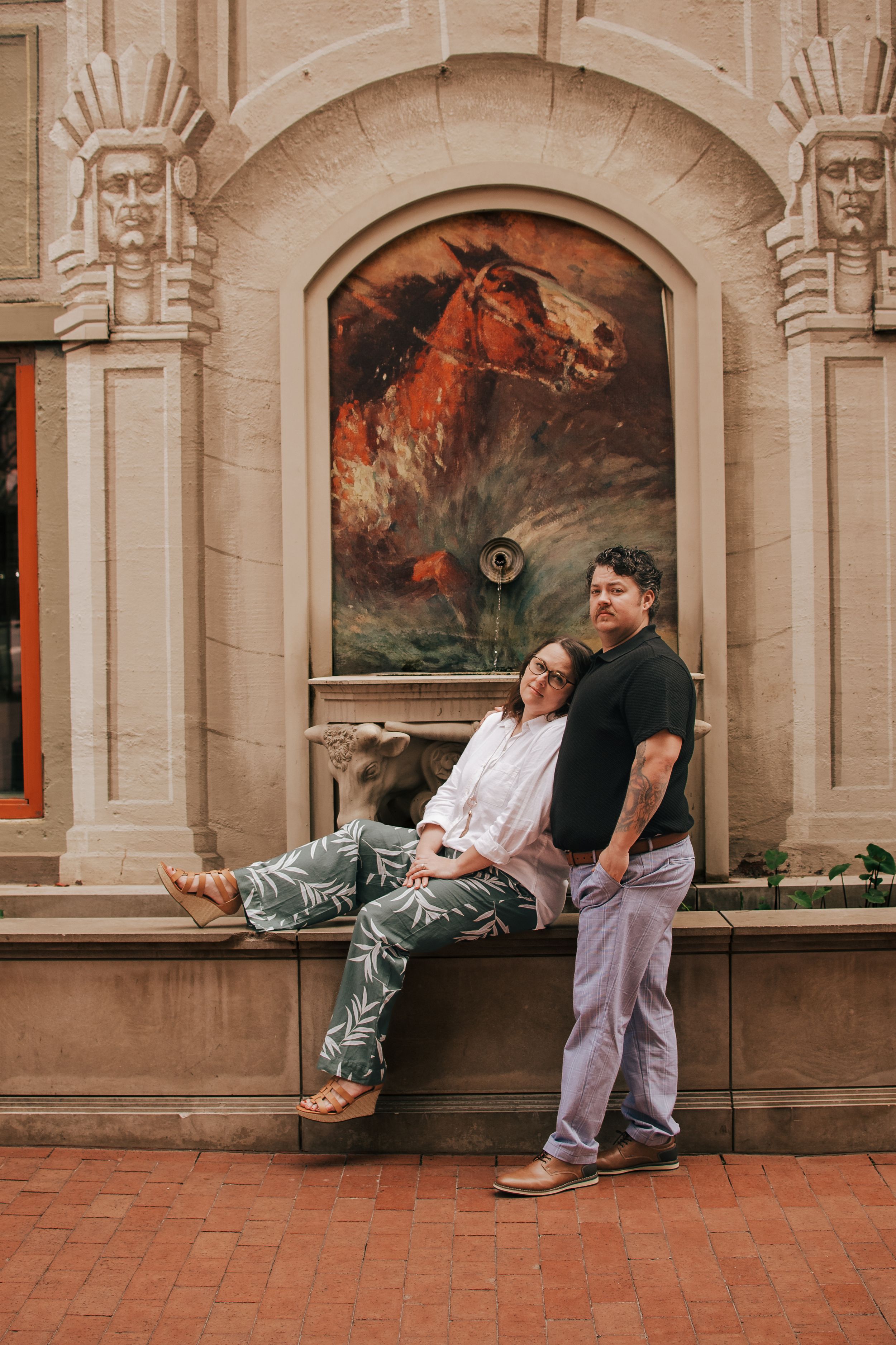 A romantic couple poses against an ornate stone building with arched architectural details and murals.