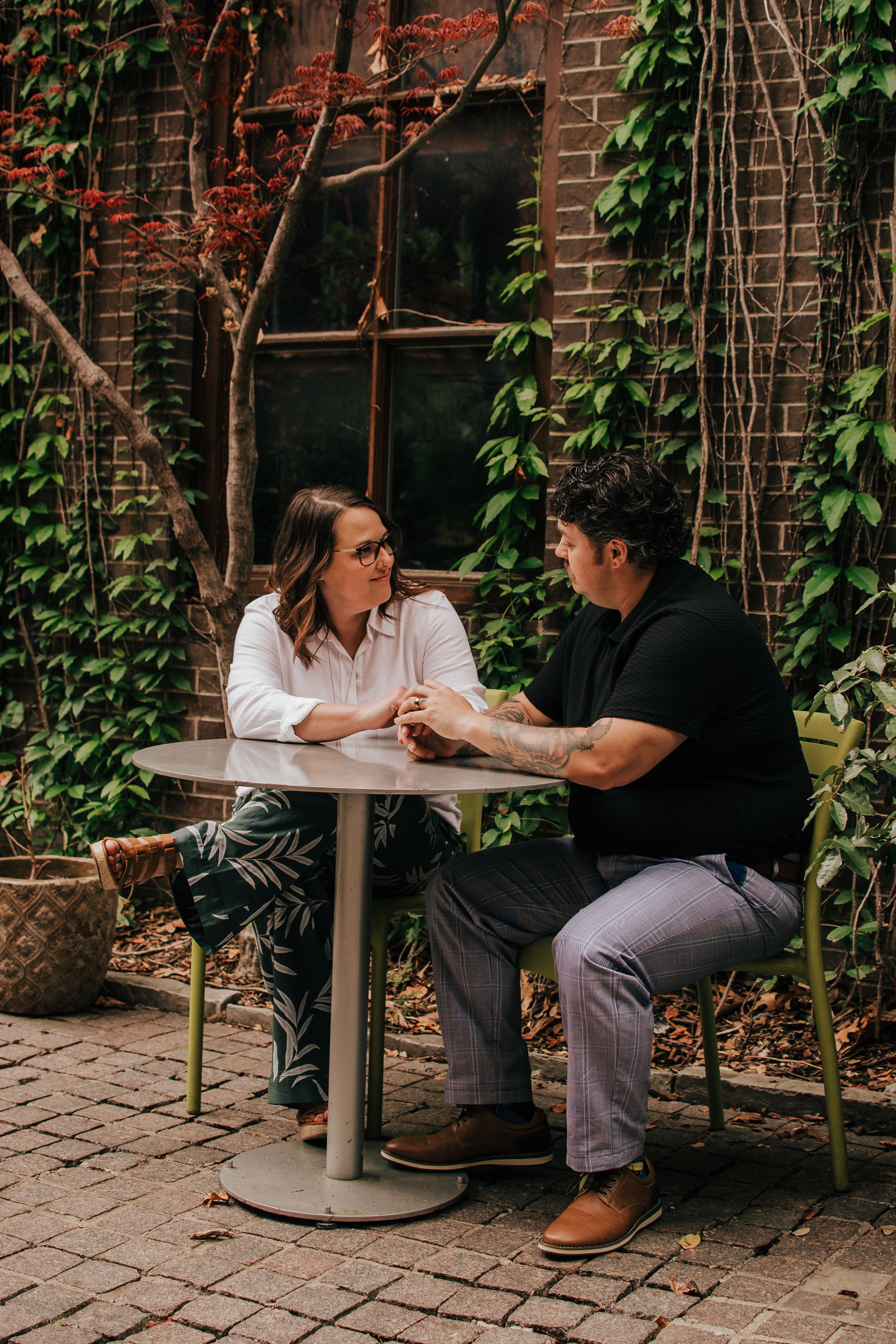 Two people engage in conversation at an outdoor table surrounded by brick walls and ivy.