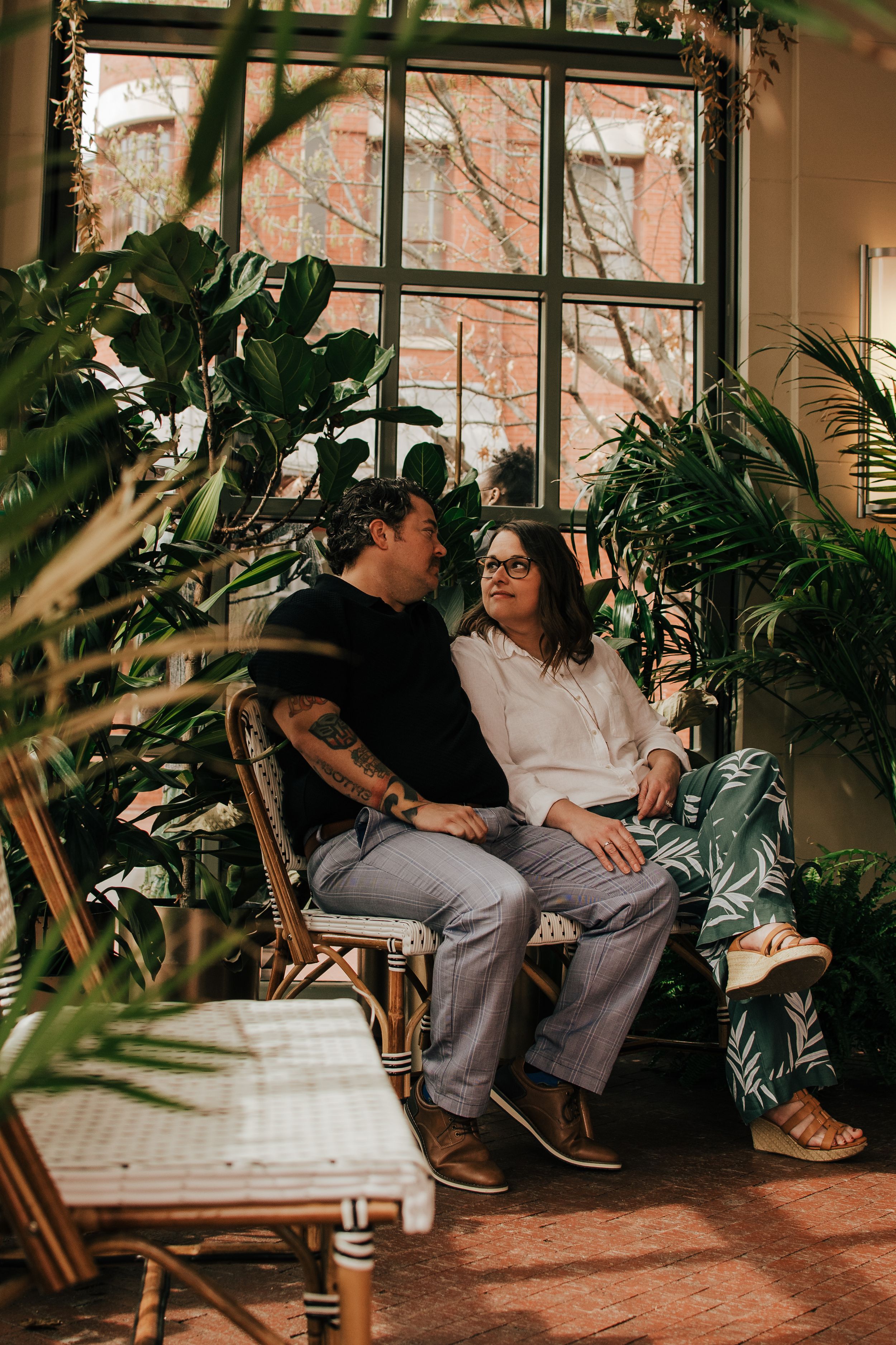 A couple relaxes on wicker furniture in a sunlit conservatory surrounded by lush tropical plants.
