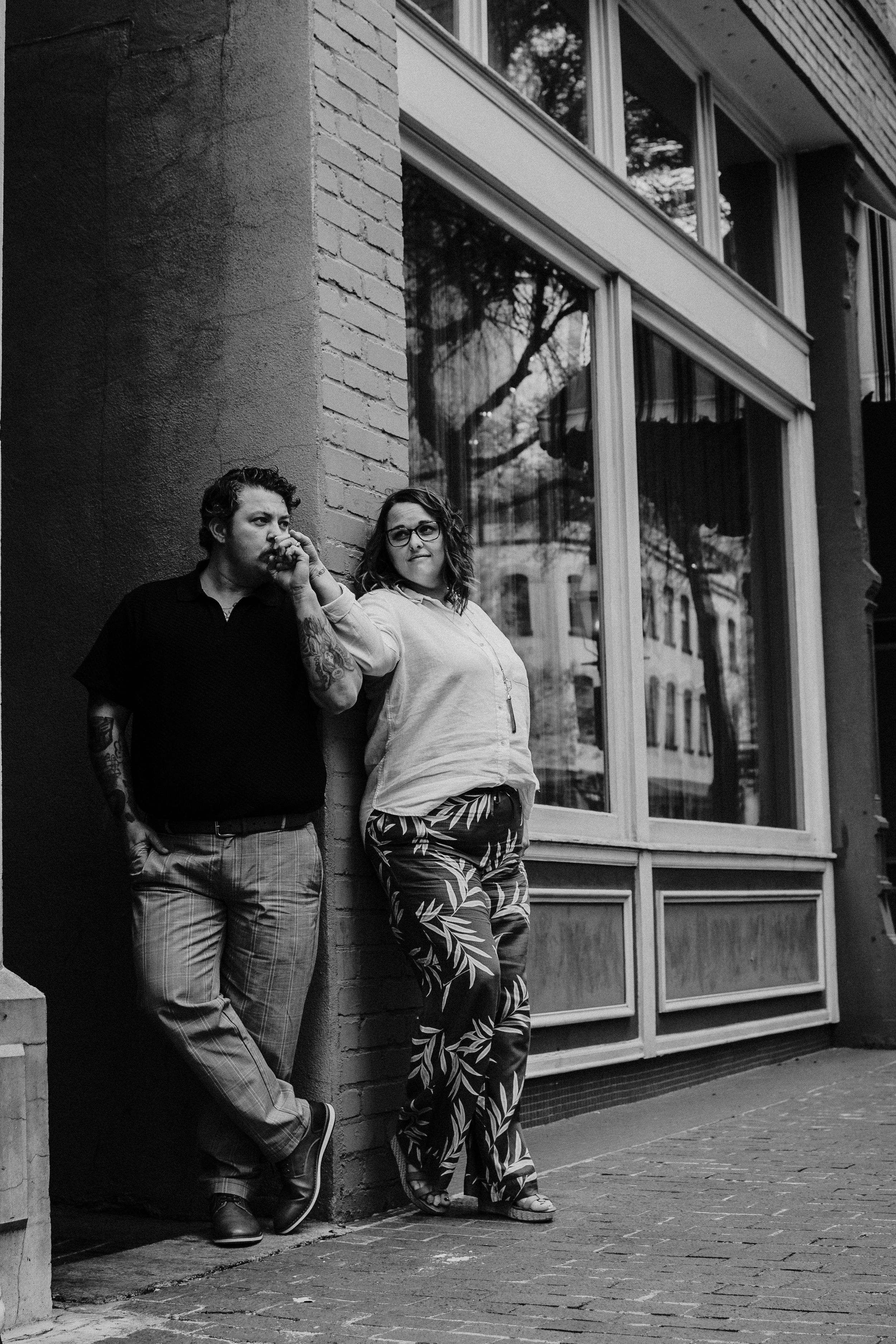 A couple leans casually against a brick storefront with large windows in an urban setting.