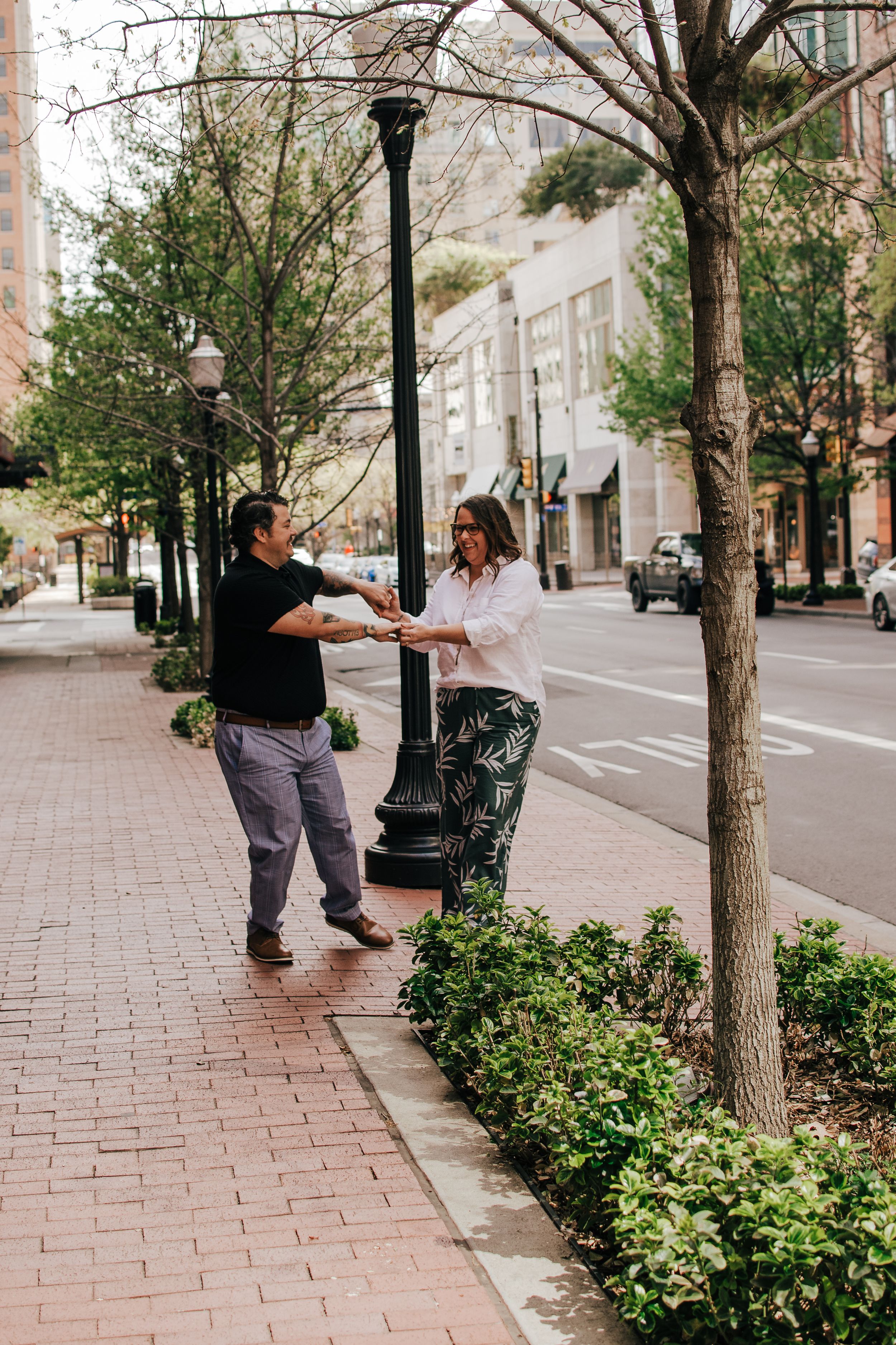 A couple shares a playful moment on a tree-lined urban sidewalk with brick pavers and lampposts.