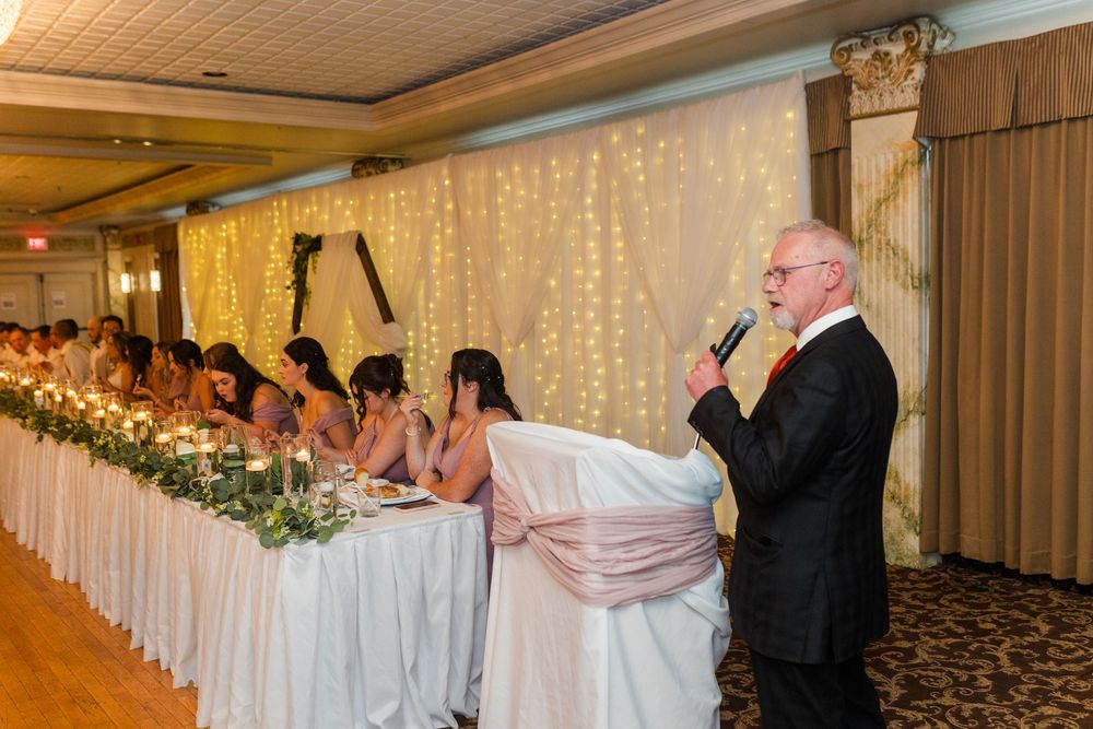 A wedding reception features couples dancing on the dance floor with a twinkle light backdrop and wooden ceiling above.
