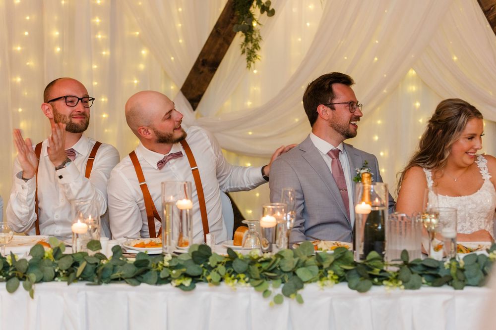 Wedding party seated at a long decorated head table with string lights and greenery garland in the background.