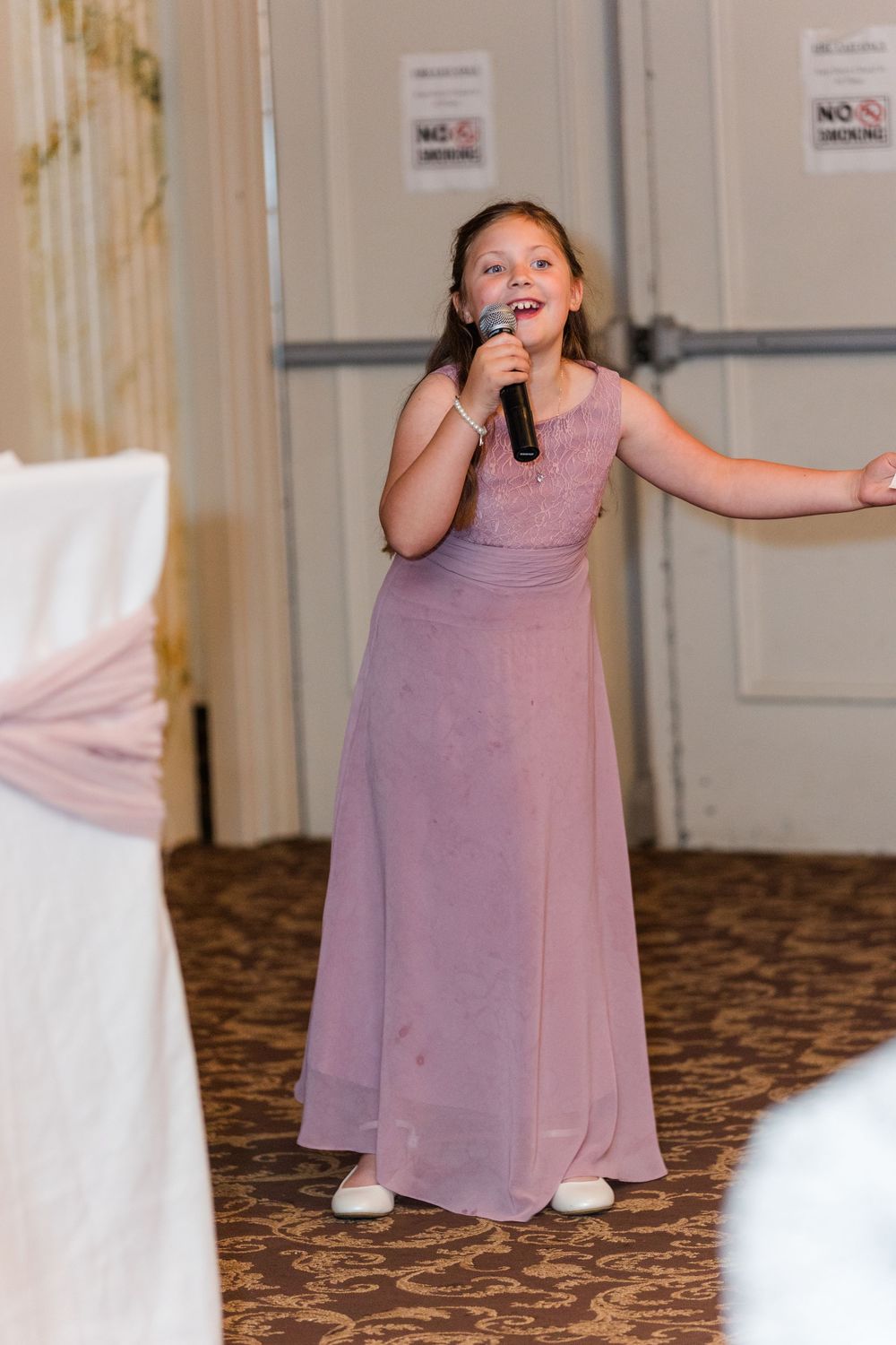 A young performer in a lavender dress sings into a microphone during what appears to be a reception or gathering.