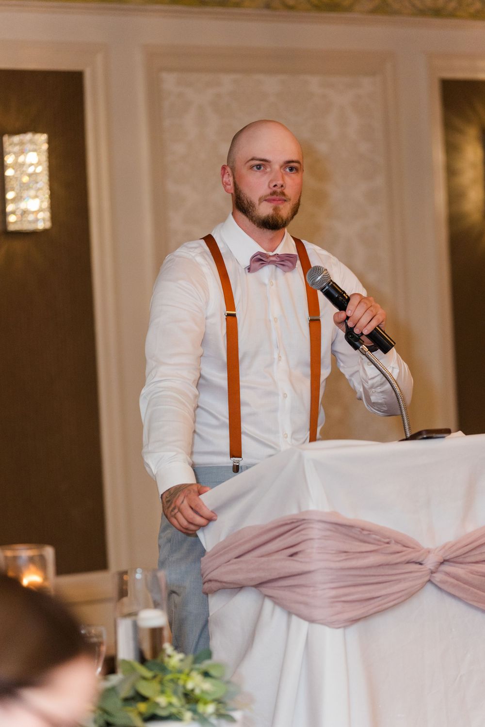 A speaker wearing orange suspenders and a white shirt makes a speech at an elegant indoor wedding reception.