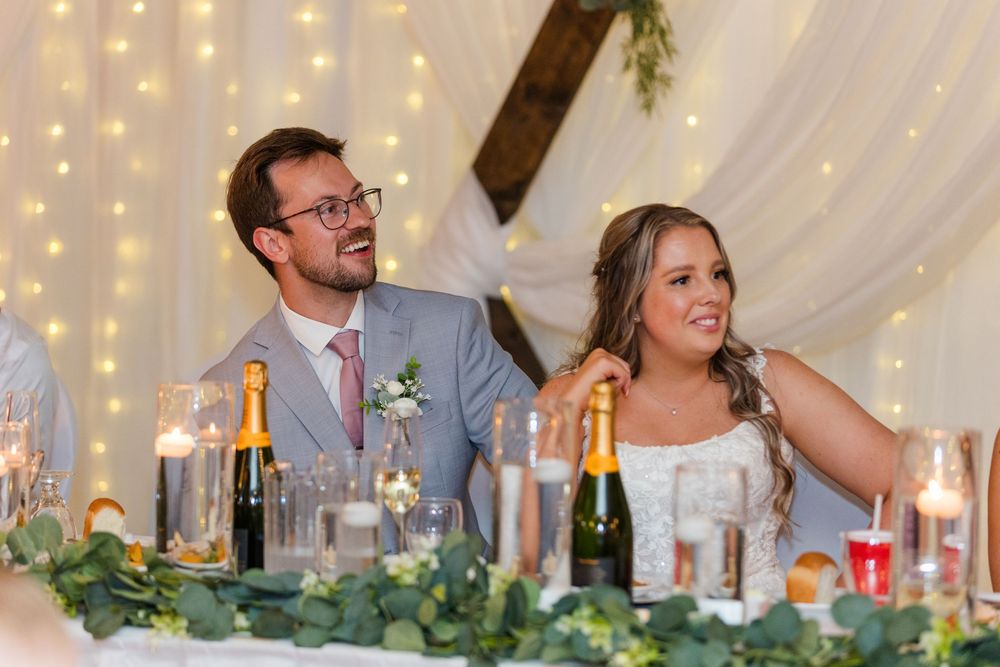 Wedding guests dining and socializing at elegant reception tables under white tent with twinkling lights and greenery decor.