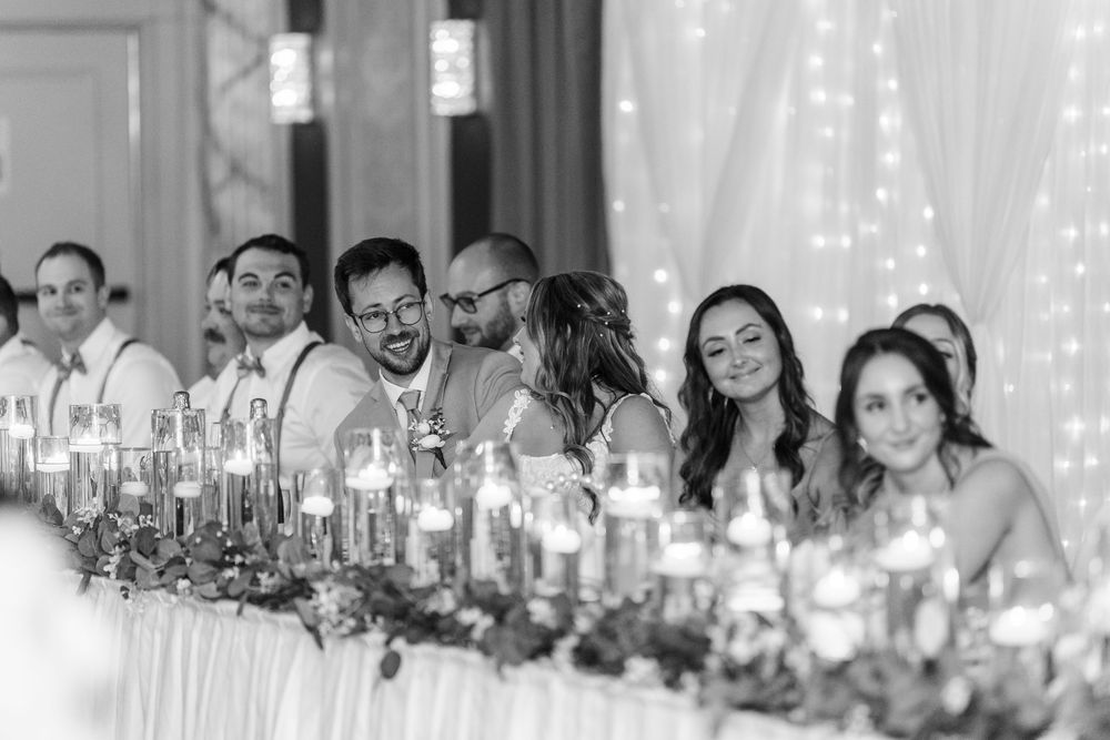 Wedding reception guests laugh and smile together at a head table decorated with glowing string lights and candles.