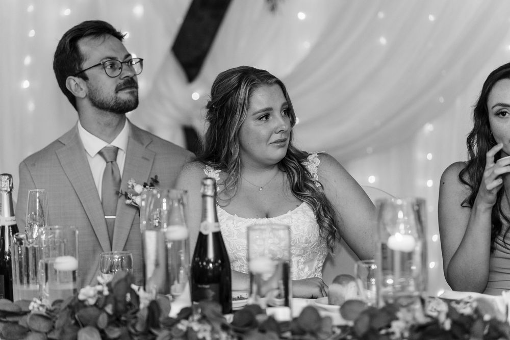 Wedding guests seated at tables under white tent during reception with wine bottles and glasses visible.