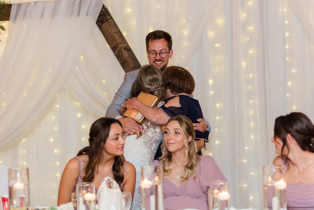 Wedding party laughing during reception toasts with twinkle lights and white drapery in background.
